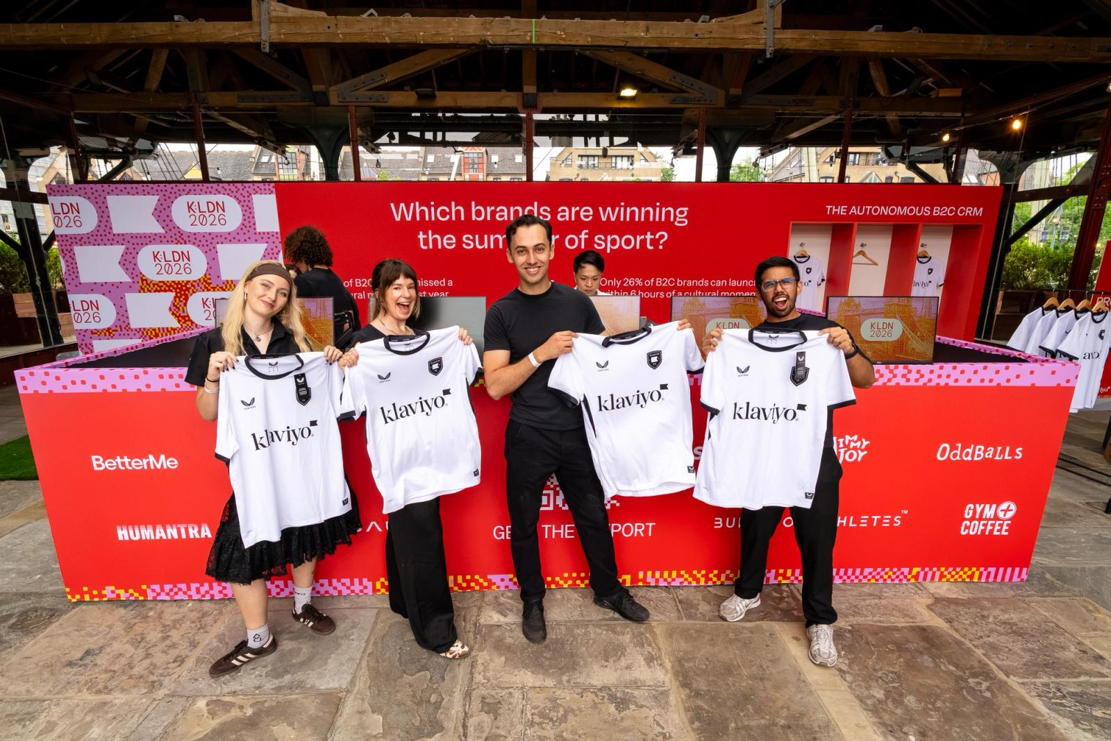 Four people hold up white sports jerseys with 'Klaviyo' printed on them, in front of a red event banner.