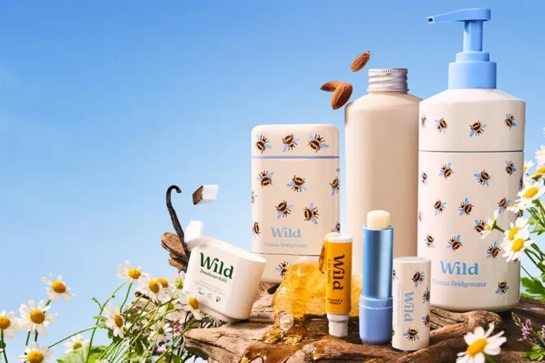 Wild Emma Bridgewater bee-patterned deodorants, lip balm, and lotion pump bottle displayed with chamomile, honeycomb, vanilla, coconut, and almonds against a blue sky.
