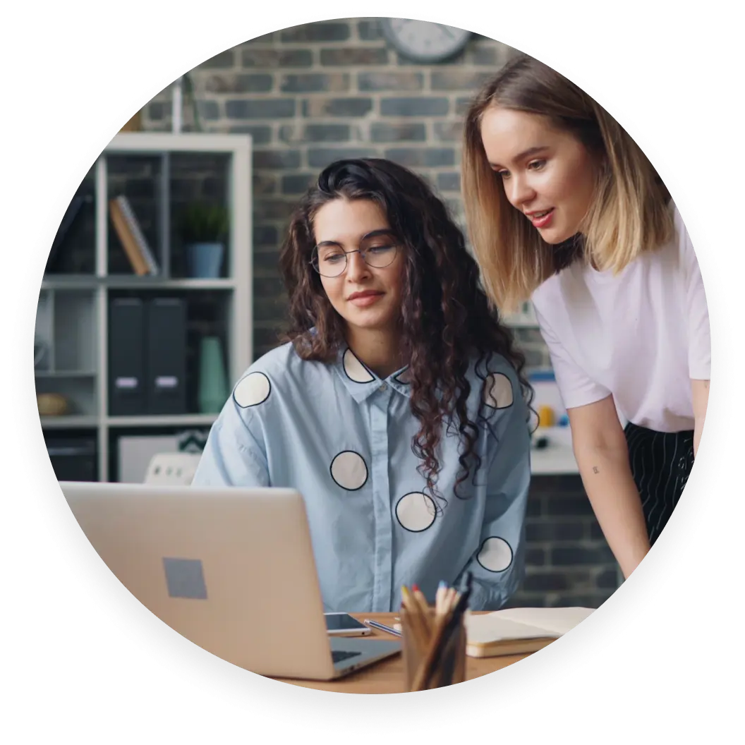 Zwei Frauen schauen auf einen Laptop-Bildschirm in einer Büroumgebung. Eine sitzt und trägt Brille und eine Polka-Dot-Bluse; die andere steht.