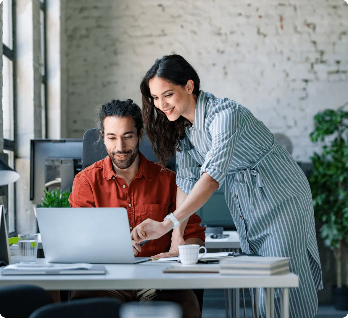 Two colleagues collaborating at a desk with a laptop, one seated and one standing, in a modern office setting.