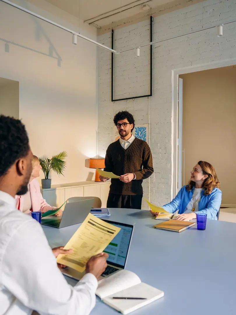 Man presents documents to colleagues gathered around a conference table with laptops and notes.