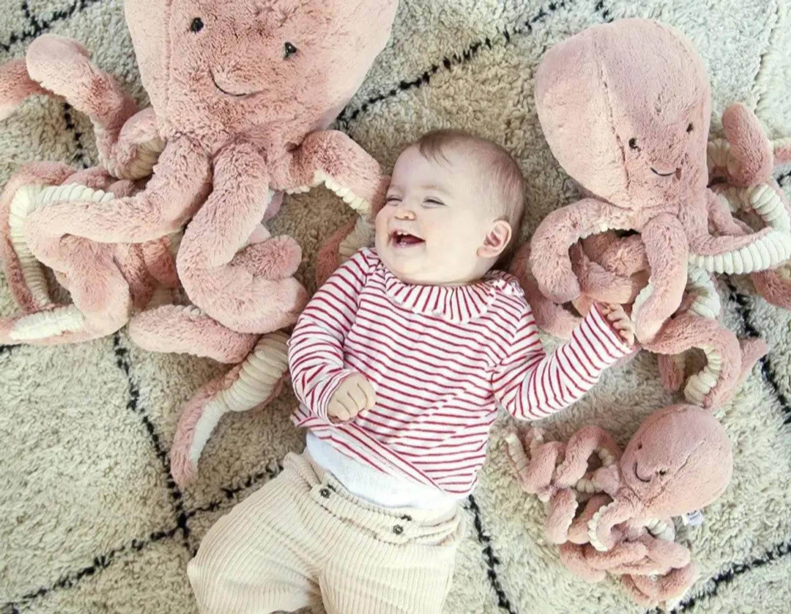 Bebé sonriendo en el suelo con varios pulpos rosas de peluche alrededor.