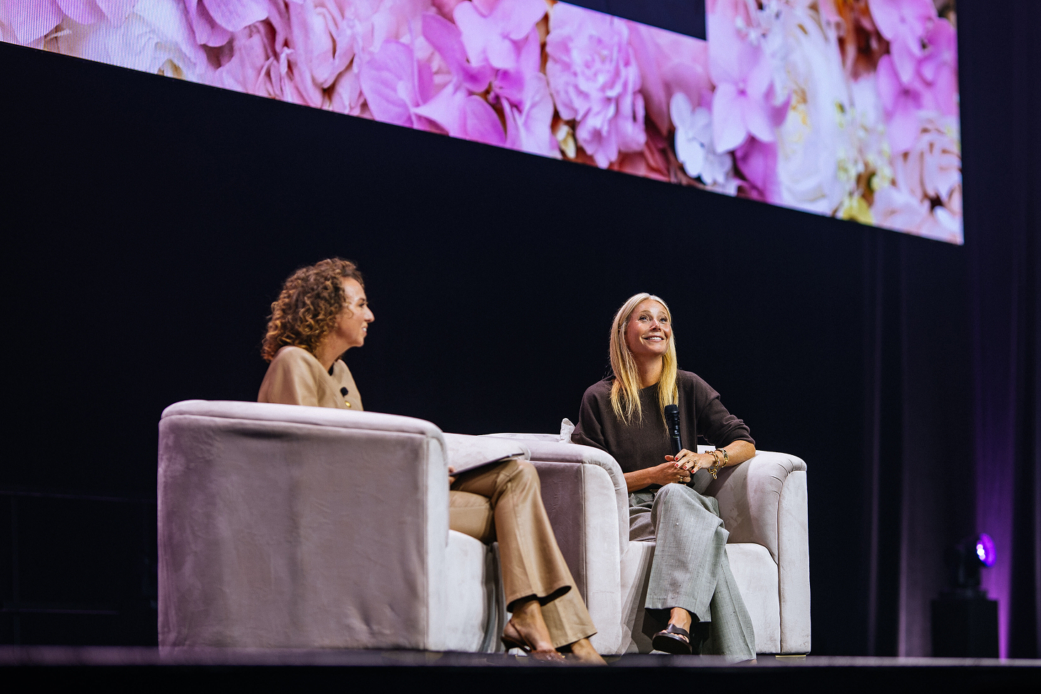 Two women seated onstage in armchairs during an interview, with one holding a microphone beneath a floral screen.