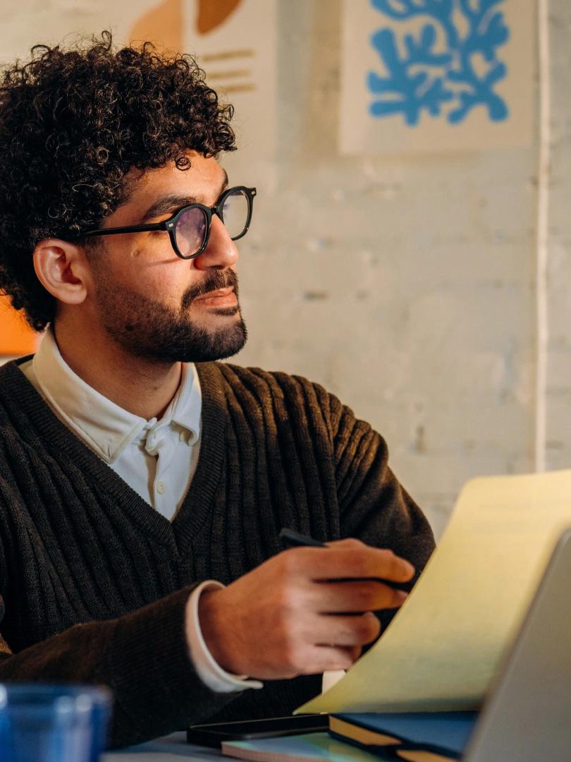 Man with curly hair and glasses holds a pen beside an open notebook and laptop.