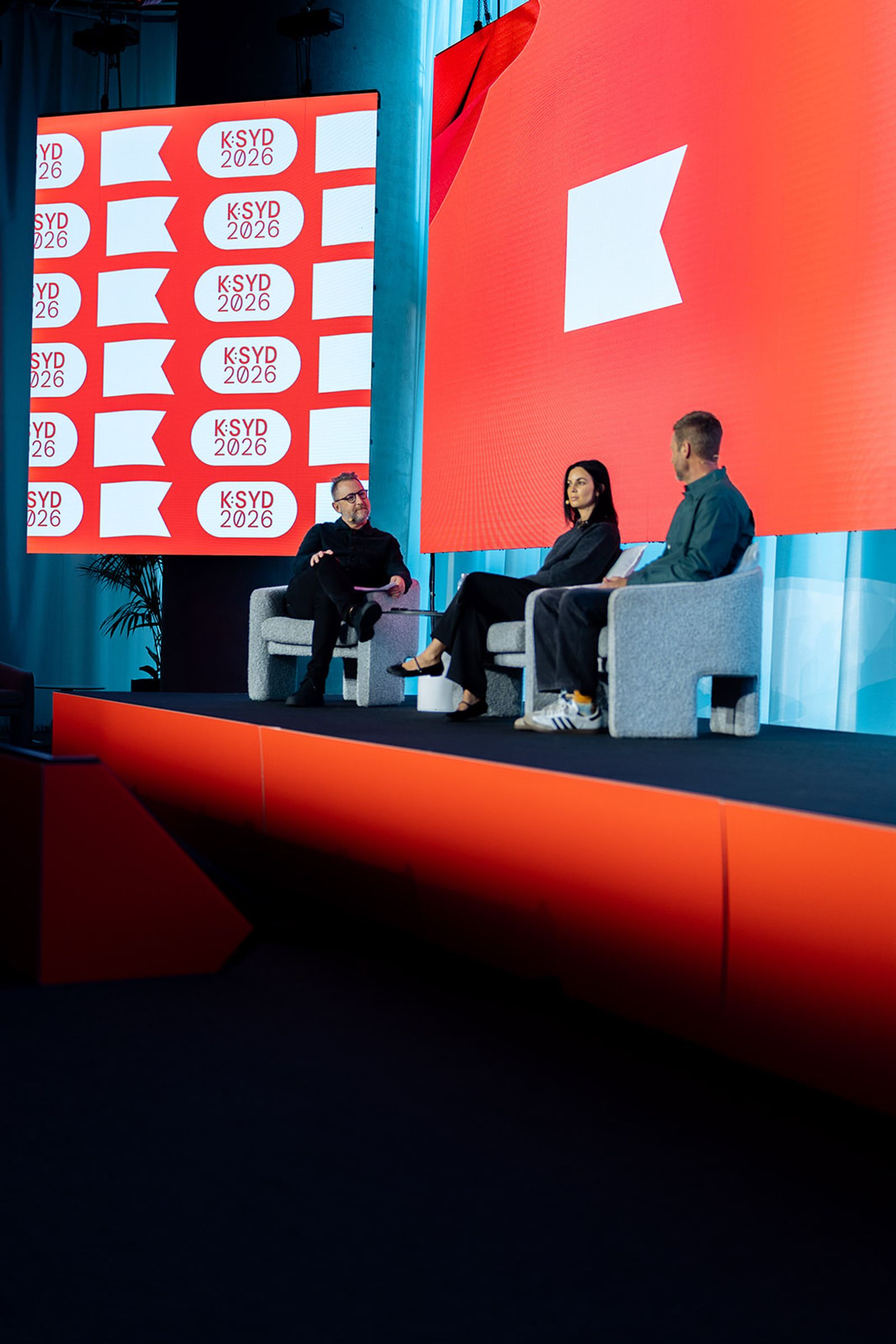Three people sit on a stage in front of large red screens displaying "KSYD 2026" and white flag graphics.