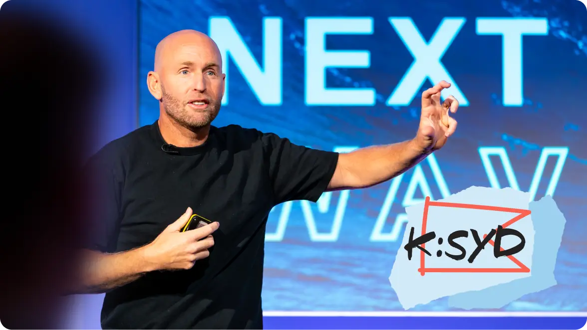 Man in a black t-shirt speaks on stage with a dynamic hand gesture, standing in front of a screen that reads "NEXT WAVE." The K:SYD logo appears in the bottom right corner.