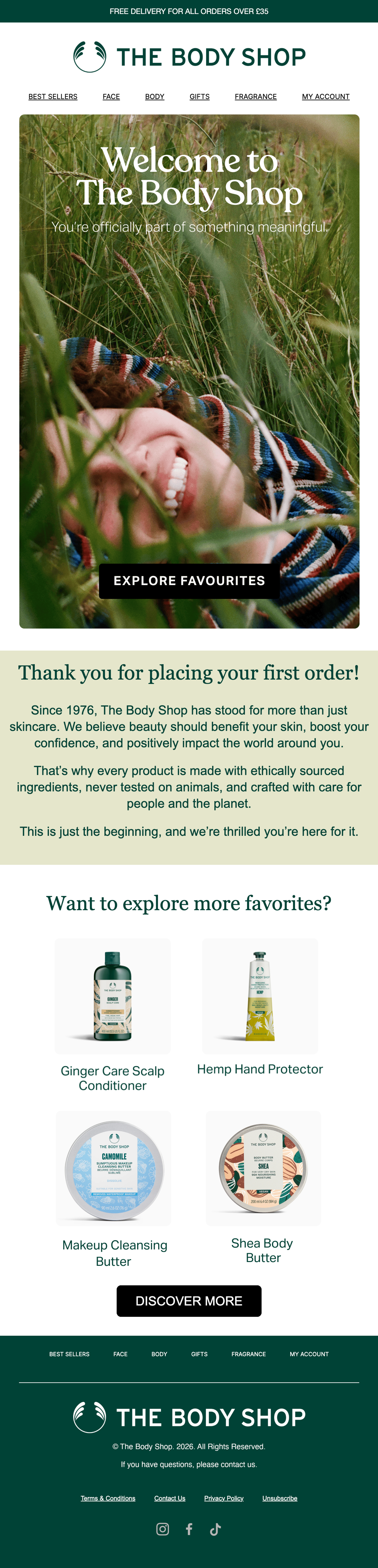 Thank-you email from The Body Shop in dark green, cream, and white. A hero photo shows a person smiling in tall grass, wrapped in a striped blanket, with the headline “Welcome to The Body Shop” and subhead “You’re officially part of something meaningful.” above an “Explore favourites” button. The heading below reads, “Thank you for placing your first order!” followed by body copy on the brand’s history, ethics, and values. The bottom half shows four product recommendations under “Want to explore more favorites?”, a “Discover more” button, and a footer with navigation, social links, and unsubscribe.