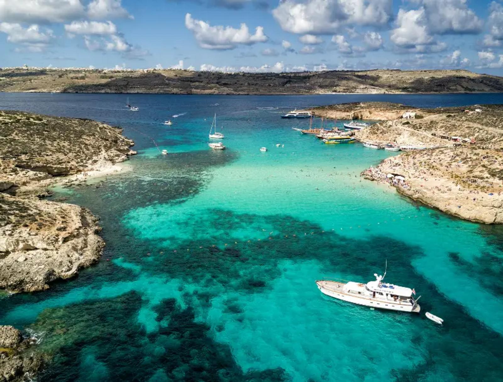 Vue aérienne du Lagon Bleu à Malte, avec ses eaux turquoise cristallines entourées de roches dorées, des bateaux amarrés et des visiteurs profitant du soleil sur la plage.