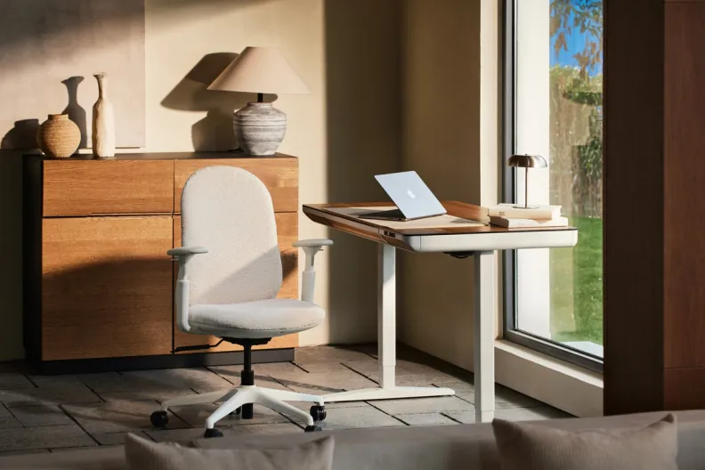 A modern home office features a white ergonomic chair, a light wood standing desk with a laptop, and a wooden credenza beside a large window looking outdoors.