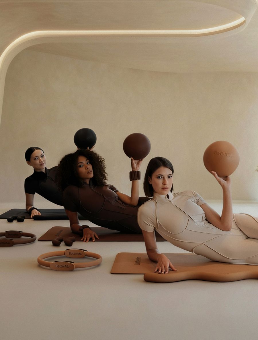 Three women in black, brown, and beige athletic jumpsuits lie on yoga mats, each holding a matching fitness ball, with exercise rings and weights on the floor.