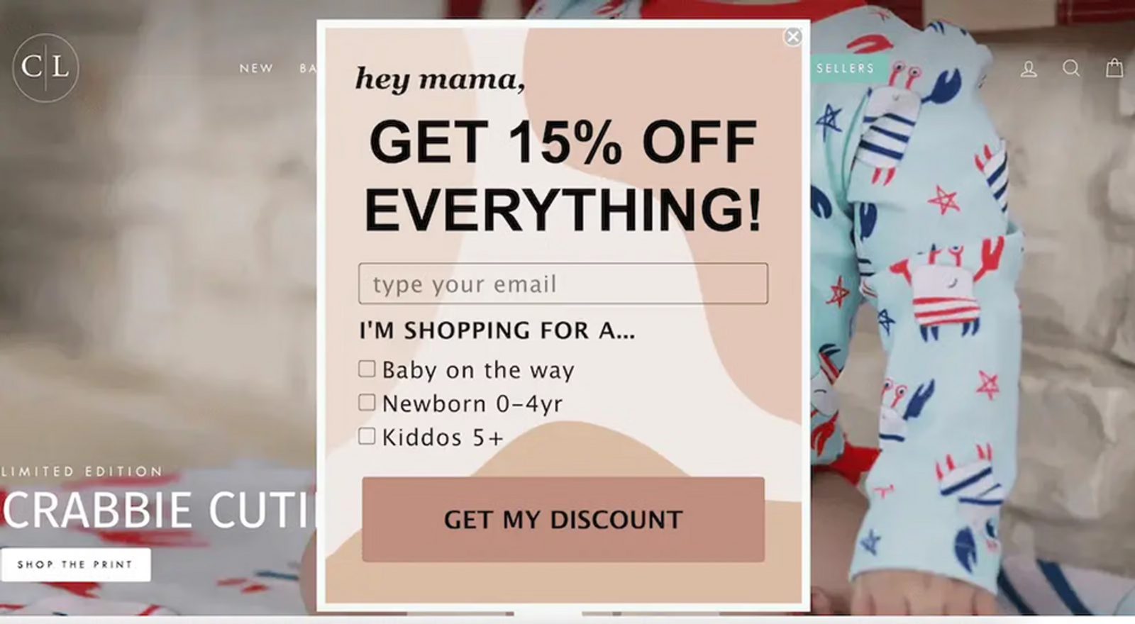 Image shows a pop-up form from Caden Lane, offering 15% off in exchange for the visitor’s email address and encouraging them to identify whether they’re shopping for a baby on the way, newborn 0-4 years old, or kids above the age of 5. In the background, on the website proper, is a partial image of a baby wearing light blue pajamas with illustrations of a cute red, white, and blue crab and hand-drawn red and blue stars.