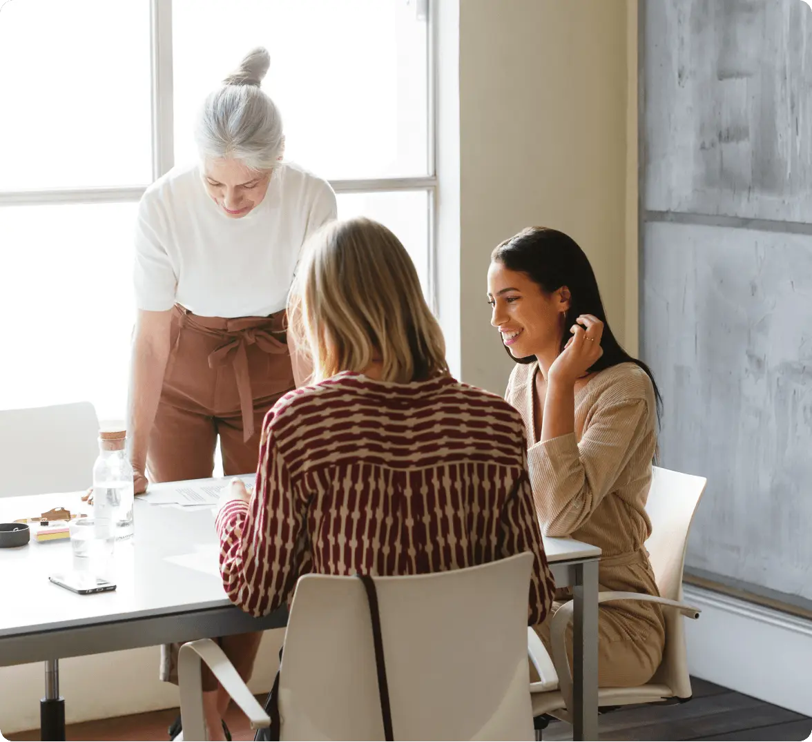 Three people having a meeting at a table, with one standing and two seated, engaged in discussion and smiling.