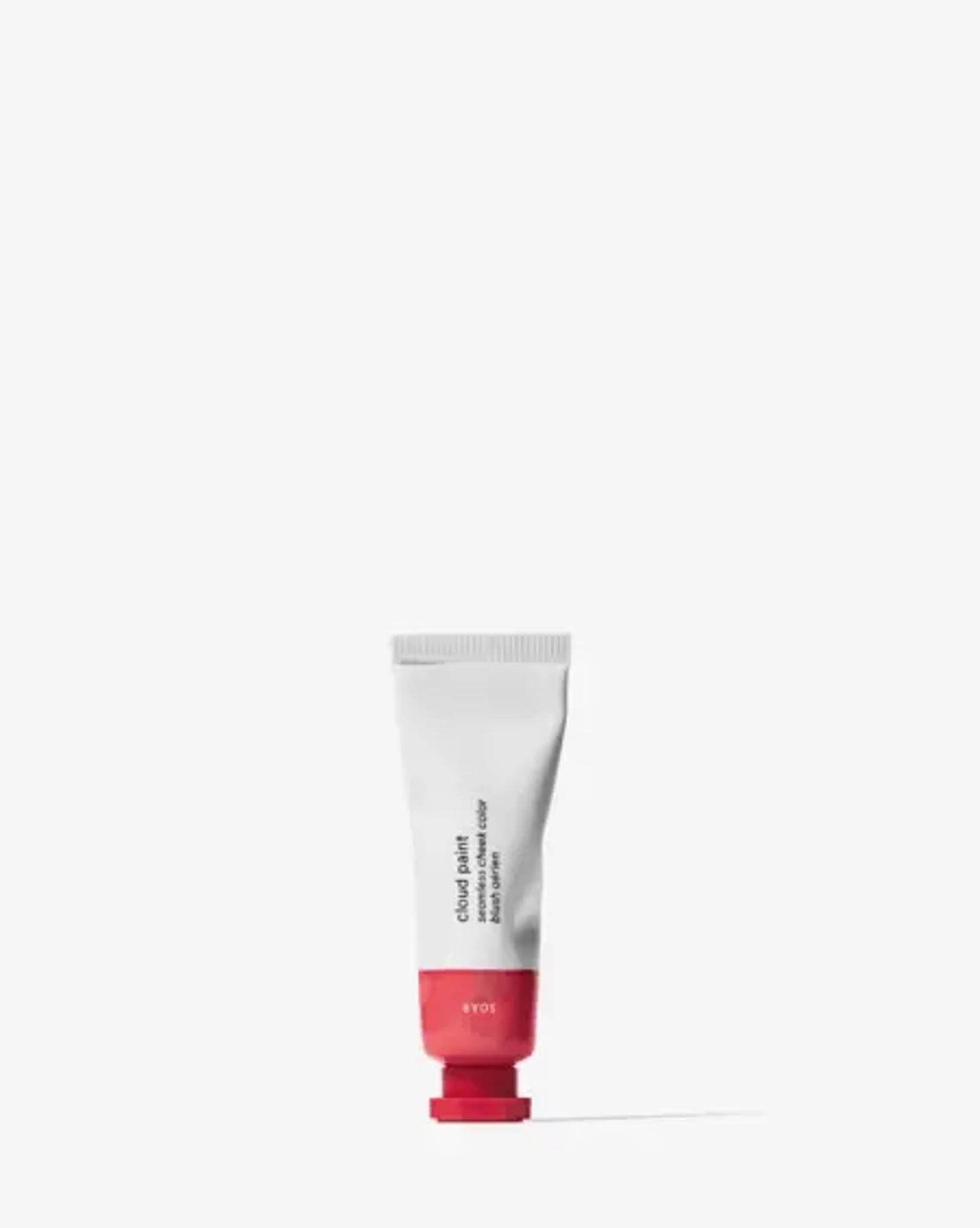 Image shows Glossier’s cloud paint product standing on its cap against a white background. The product is a white bottle with black text and a red end cap.