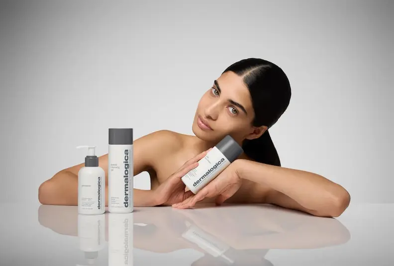 A woman with bare shoulders posing with three Dermalogica skincare products on a reflective surface.