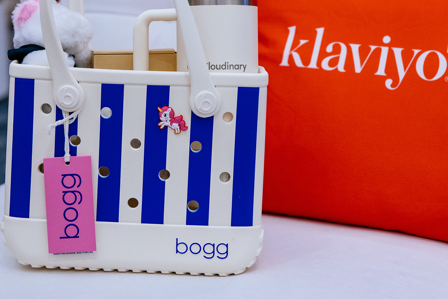Blue-and-white striped Bogg tote with a unicorn charm, tumbler and plush toy beside a red Klaviyo backdrop.