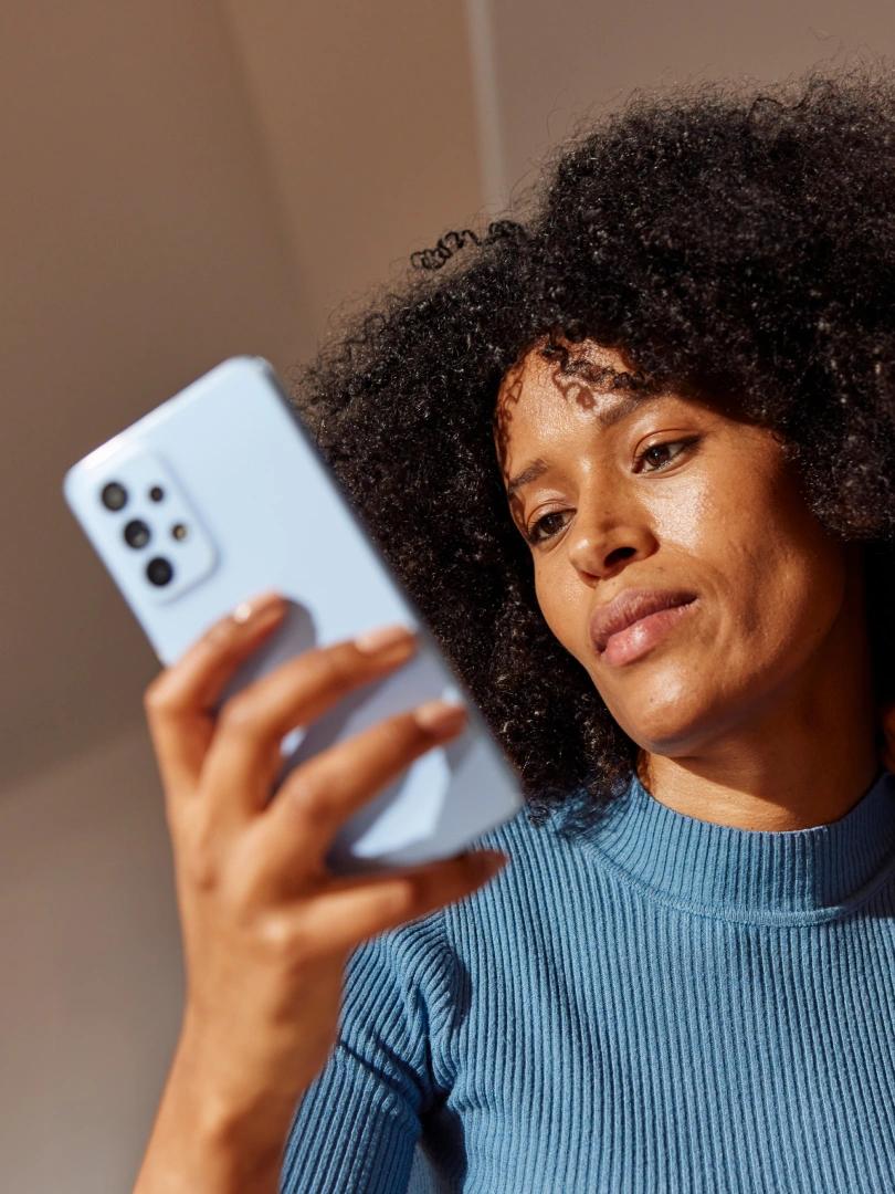Woman with curly hair looks at a light blue smartphone in warm sunlight.
