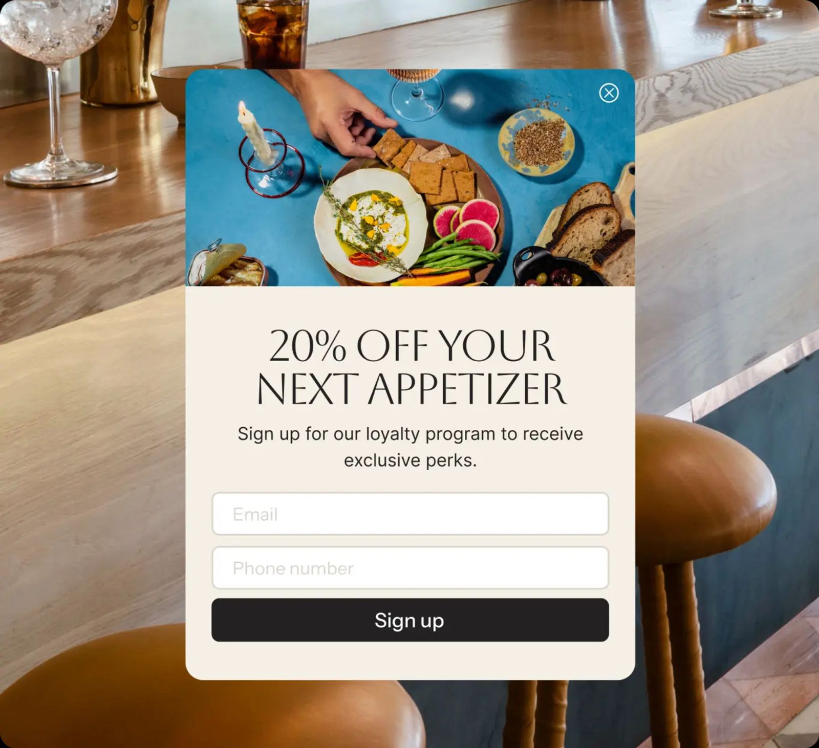 Image shows a pop-up sign-up form. A photograph at the top shows a spread of dips, crackers, radishes, green beans, olives, and bread on a blue table. Below, a headline reads “20% OFF YOUR NEXT APPETIZER” and body copy reads “Sign up for our loyalty program to receive exclusive perks.” Two empty fields follow, labelled “Email” and “Phone number,” above a black “Sign up” button.