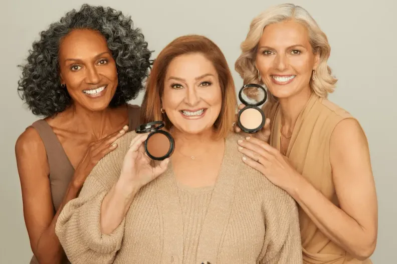 Three smiling women of diverse ages and ethnicities holding open makeup compacts with varying shades.