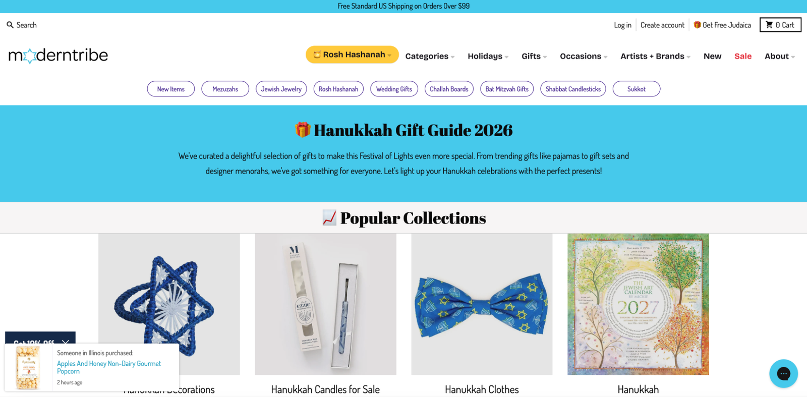 Screenshot of the ModernTribe website's Hanukkah Gift Guide 2026 landing page. The top navigation bar includes search, a Rosh Hashanah dropdown, and menu links for categories, holidays, gifts, occasions, artists + brands, new, sale, and about, with a row of category buttons below for mezuzahs, Jewish jewelry, Rosh Hashanah, wedding gifts, challah boards, bat mitzvah gifts, shabbat candlesticks, and sukkot. A blue banner reads, "Hanukkah Gift Guide 2026," with text describing a curated selection of gifts including pajamas, gift sets, and designer menorahs. Below, a "popular collections" section shows four product images: a blue crocheted Star of David ornament, a boxed Hanukkah candle set, a blue bow tie printed with menorahs and Stars of David, and a 2027 Jewish art calendar featuring illustrated trees. A small pop-up in the bottom left shows a recent purchase notification for "apples and honey non-dairy gourmet popcorn."
