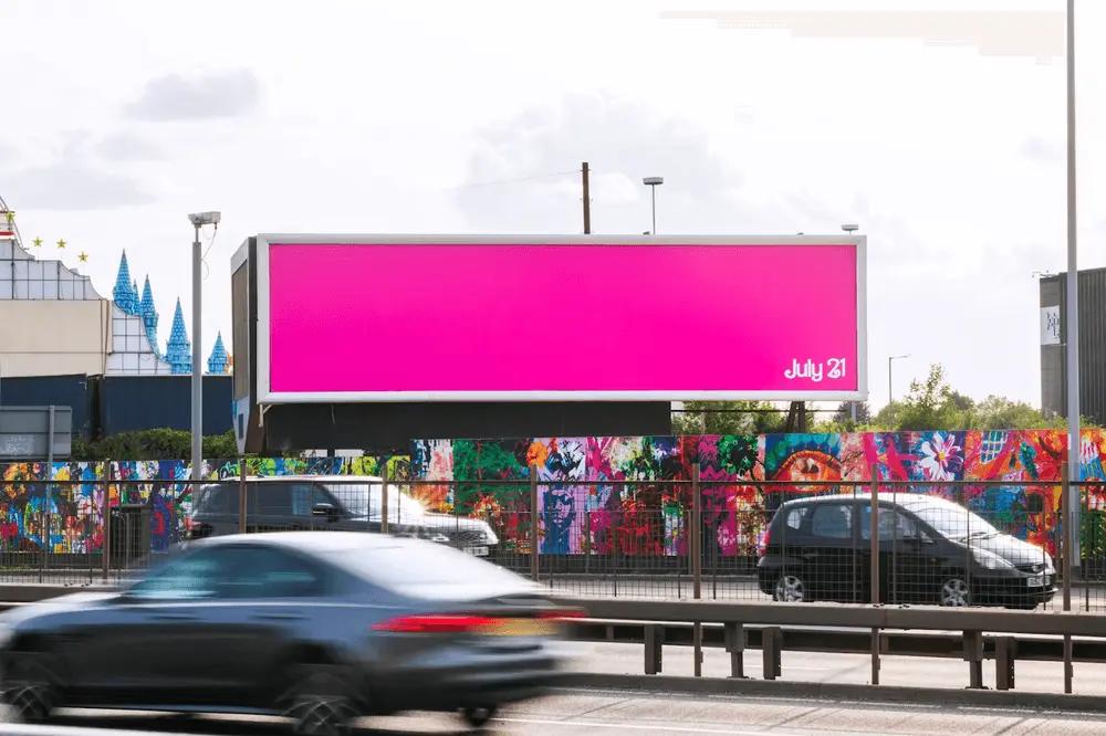 Image shows a Barbie Pink billboard with a multi-colored mural beneath it. 3 cars pass by, blurred to indicate speed. The date July 21 in the bottom-right corner in white is the only graphic element on the billboard besides the color.