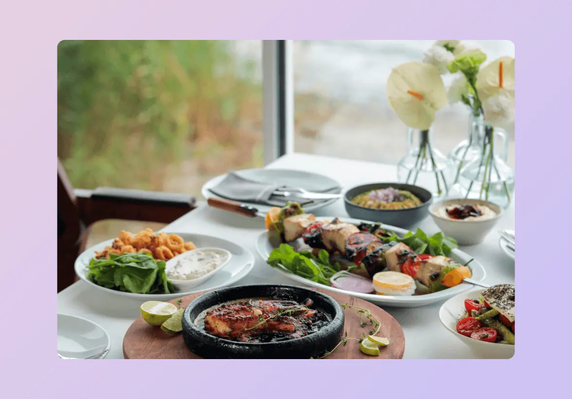 A table set with various dishes, including grilled meat, skewers, salads, and sauces, next to a window with vases of flowers.