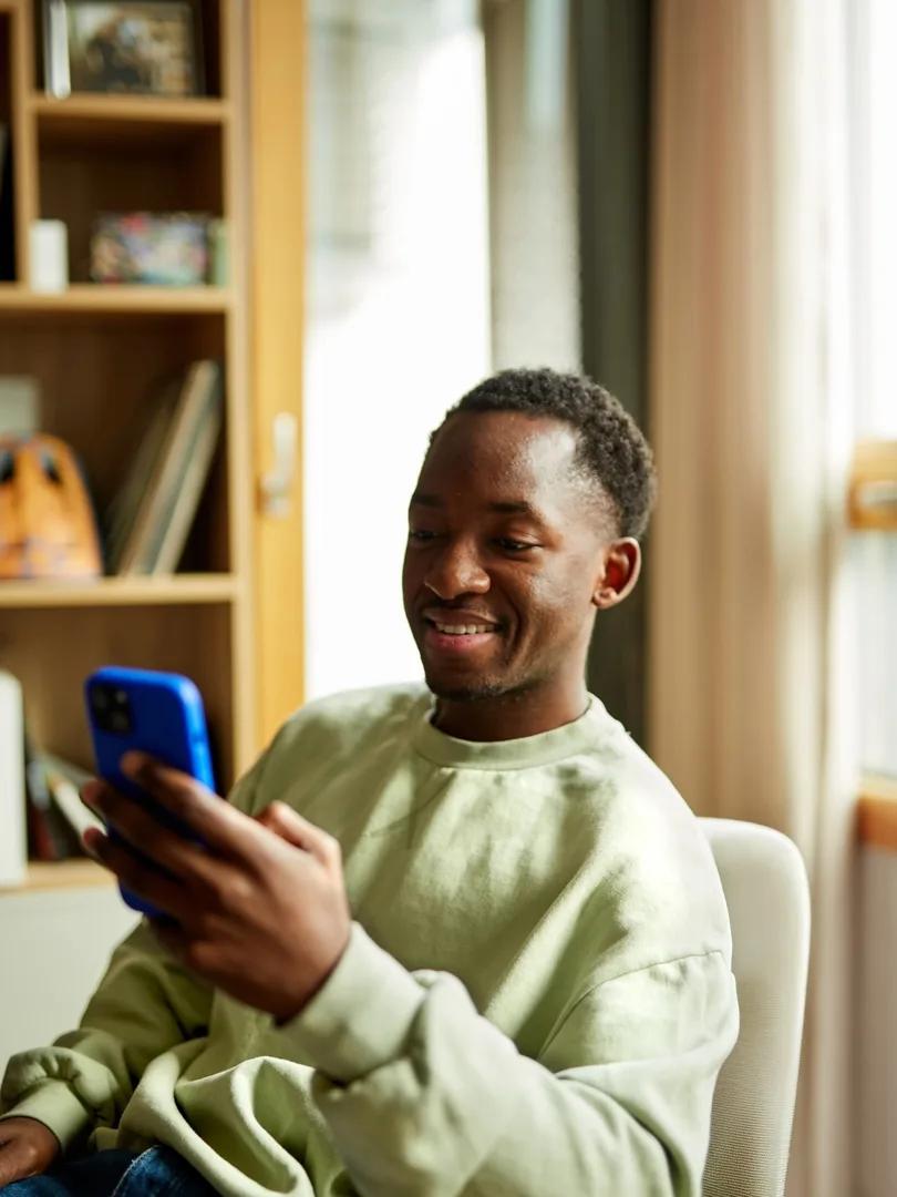 Smiling young man seated indoors and looking at a blue smartphone.