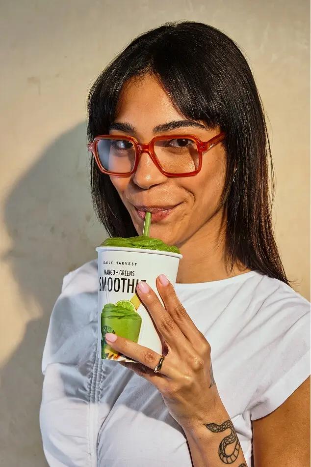 Woman in red glasses sips a green mango smoothie from a cup.