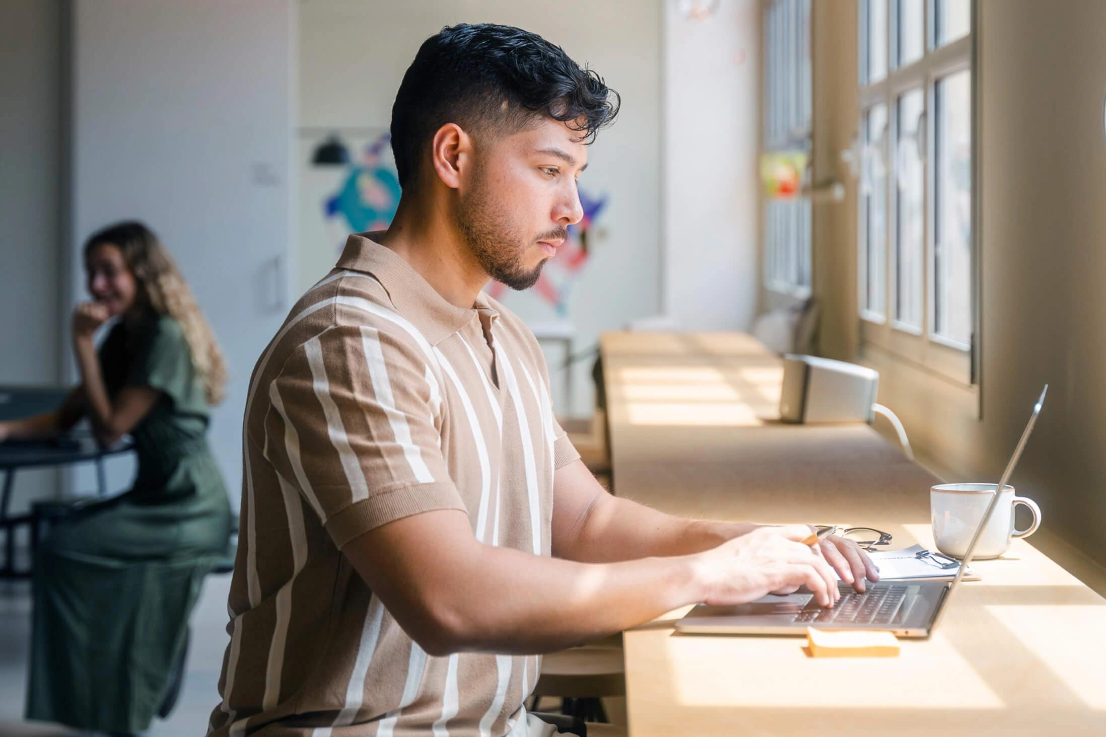 Young man works on a laptop at a sunlit counter, with a coffee cup beside him.