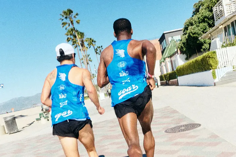 Rear view of two men running in blue tank tops on a sunny boardwalk with palm trees.