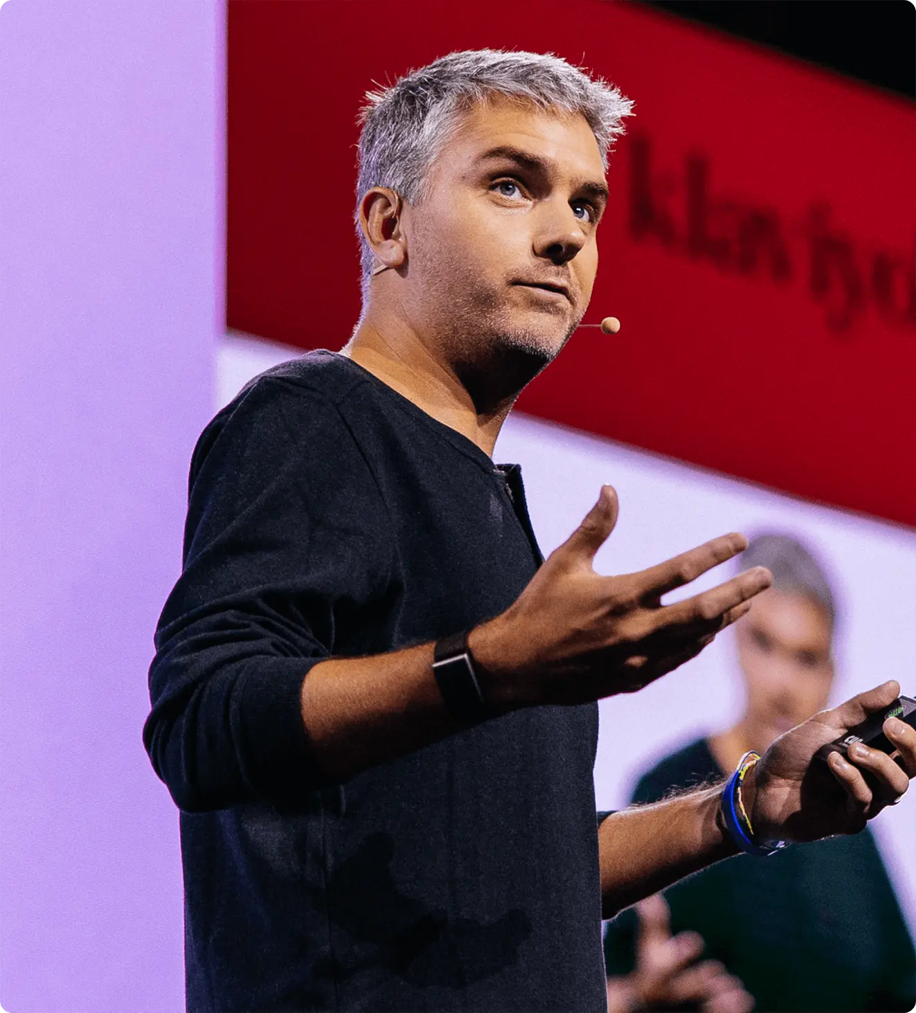 A person with a headset microphone gestures while speaking on stage, with a blurred presentation screen in the background.