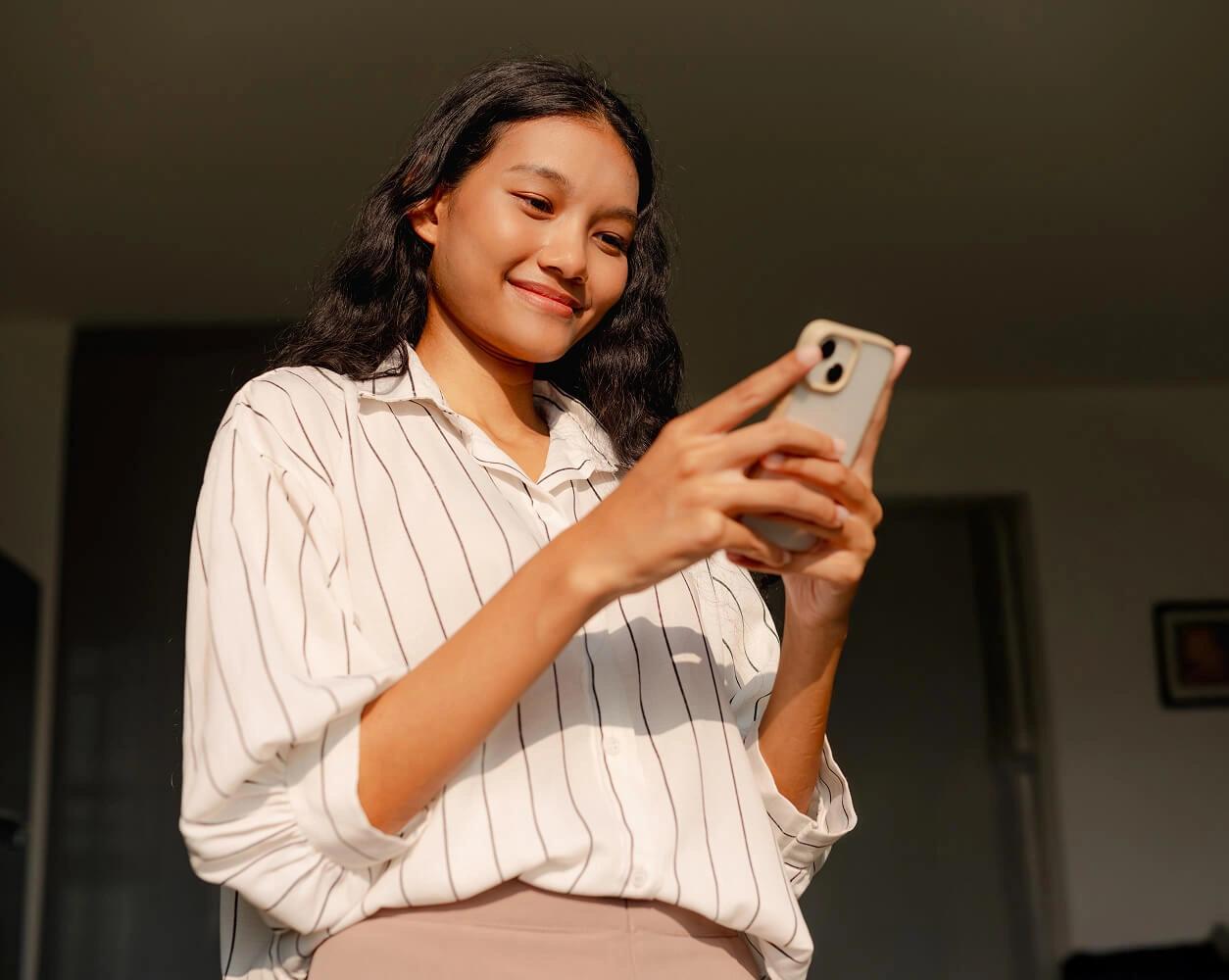 Smiling young woman using a smartphone in warm sunlight indoors