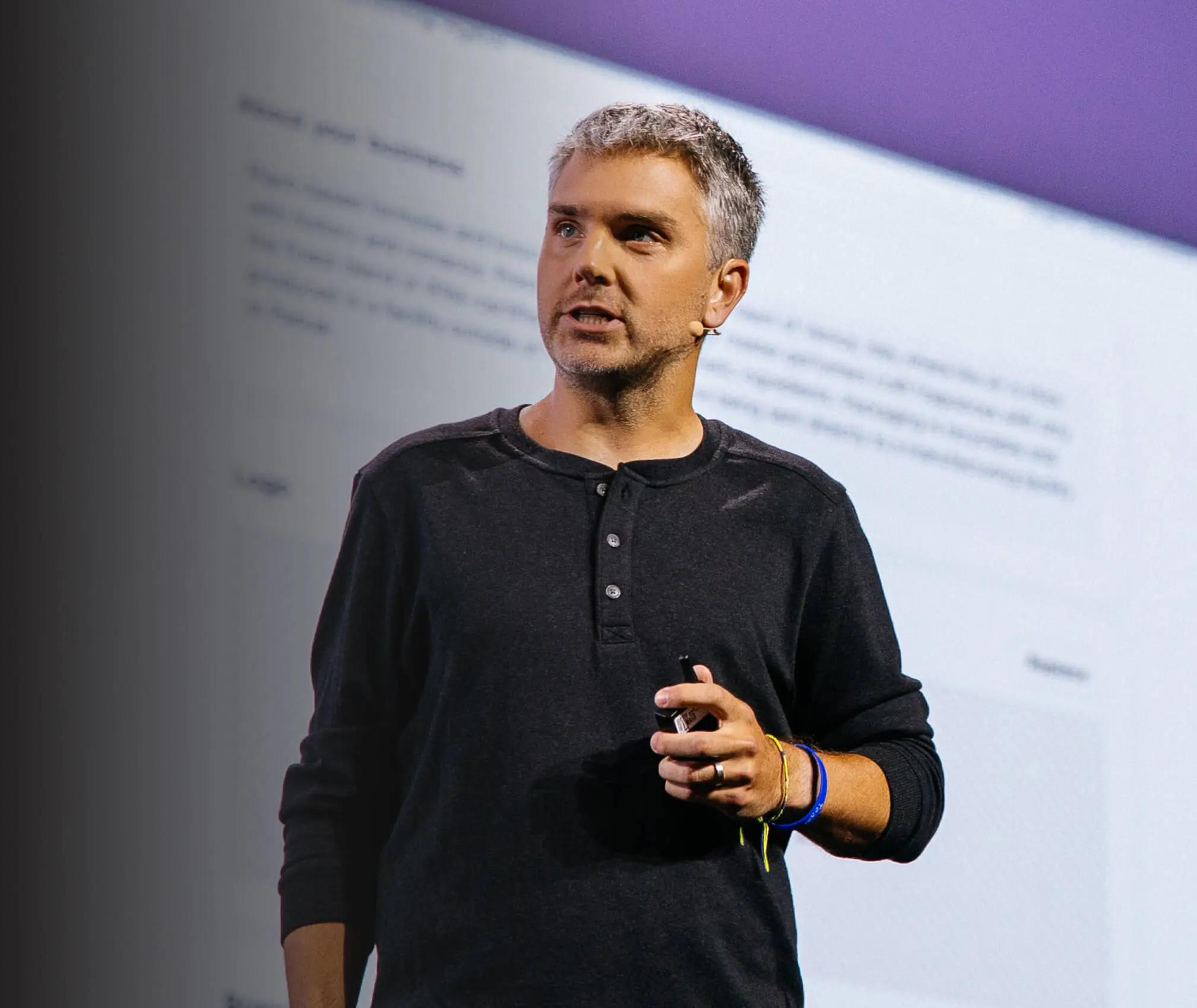 A man with short gray hair speaks on stage, holding a microphone. He wears a dark henley shirt and wristbands.