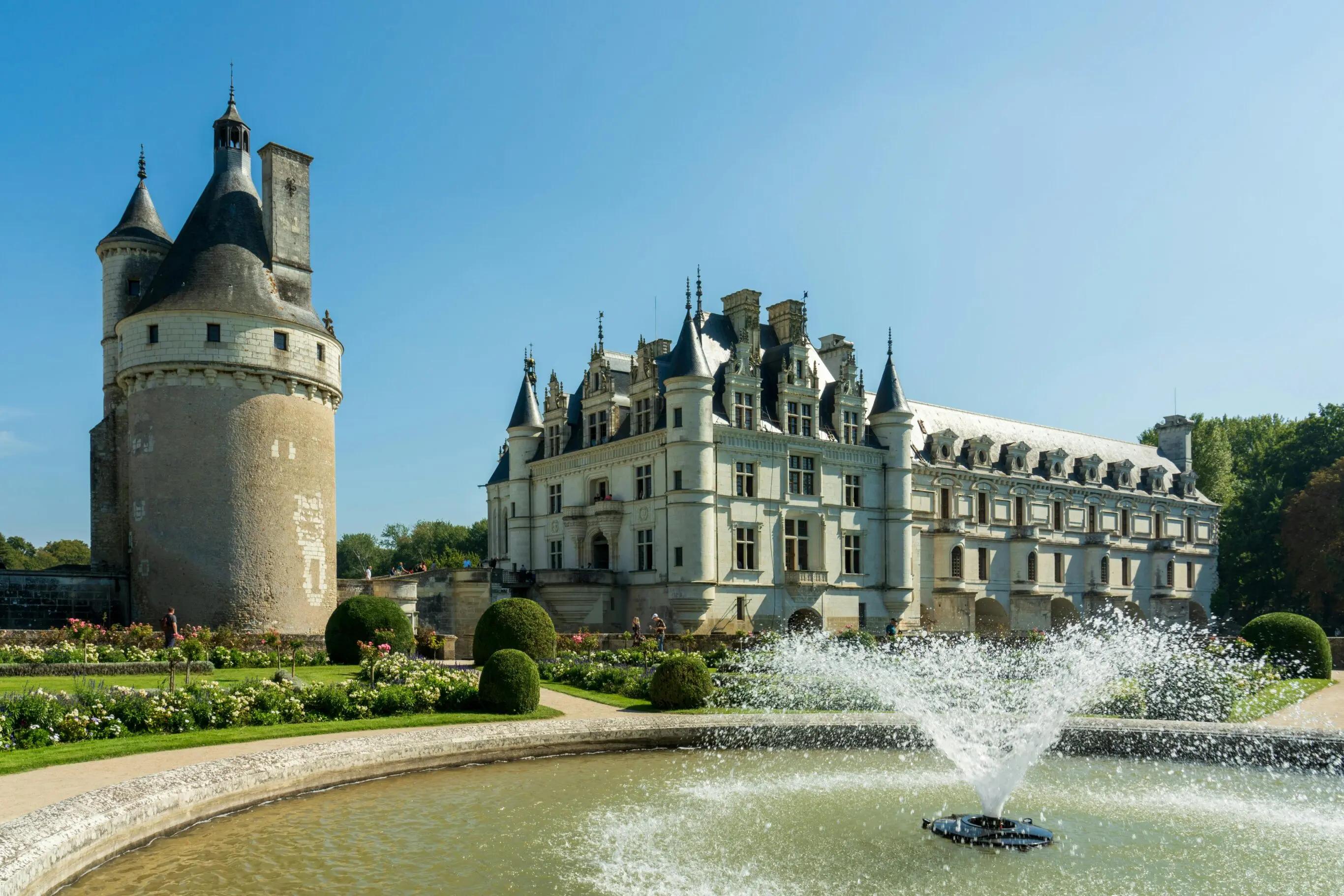  Château de Chenonceau en France, entouré de jardins fleuris et d'une fontaine en premier plan, sous un ciel bleu clair.