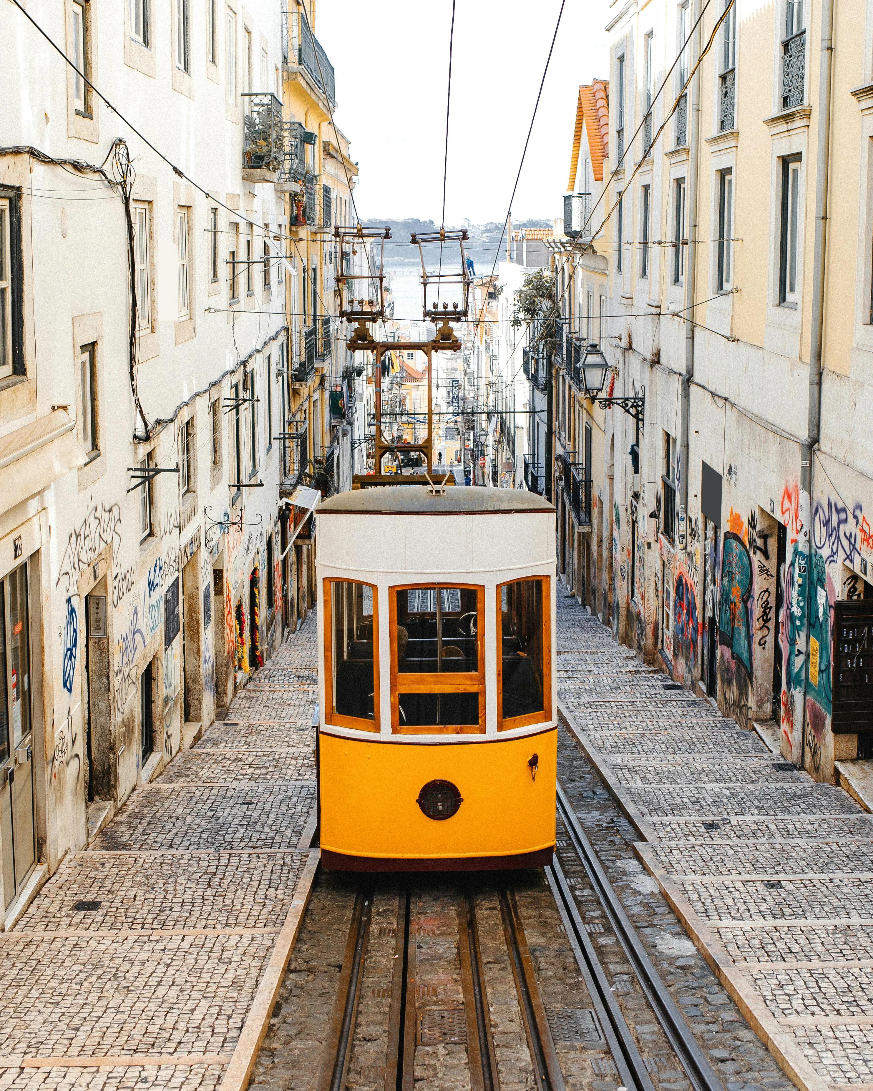 Un tramway jaune emblématique de Lisbonne descendant une rue pavée escarpée, entourée de bâtiments aux façades couvertes de graffitis et de balcons en fer forgé, avec une vue sur le Tage en arrière-plan.