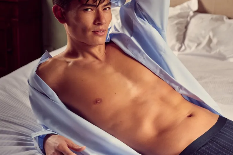 A man with an open blue shirt and striped underwear lies on a bed, looking at the viewer.