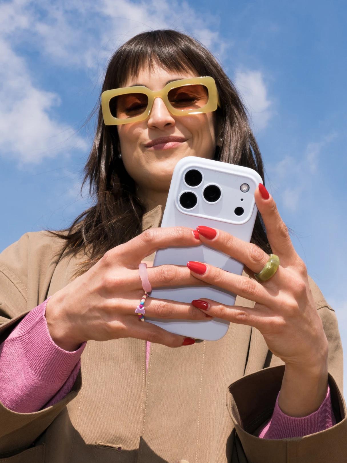Woman in oversized sunglasses uses a smartphone outdoors beneath a blue sky.