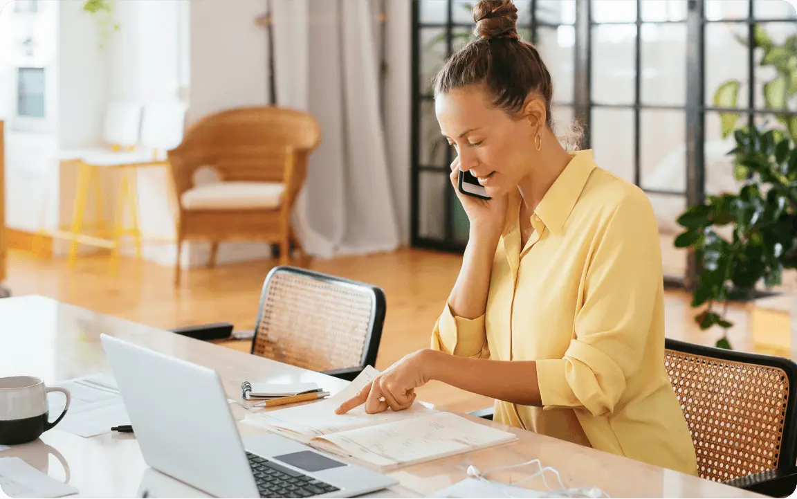 Donna al telefono seduta a una scrivania con un laptop e documenti. Indossa una camicia gialla e sembra concentrata.
