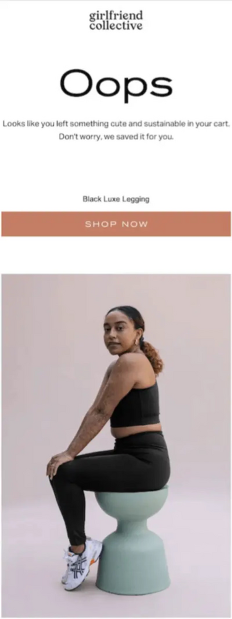 An email by Girlfriend Collective that opens with their logo in black text at the top. Underneath, a large header reads, “Oops” in black text against a white background. Underneath the text is a taupe CTA button that reads “Shop Now” in white text. Underneath that block is an image of a dark-skinned person with light and dark brown hair pulled back in a low ponytail sitting on a green bucket chair wearing a cropped black tank top and black leggings. They’re sitting sideways to the camera with their face turned toward the camera.