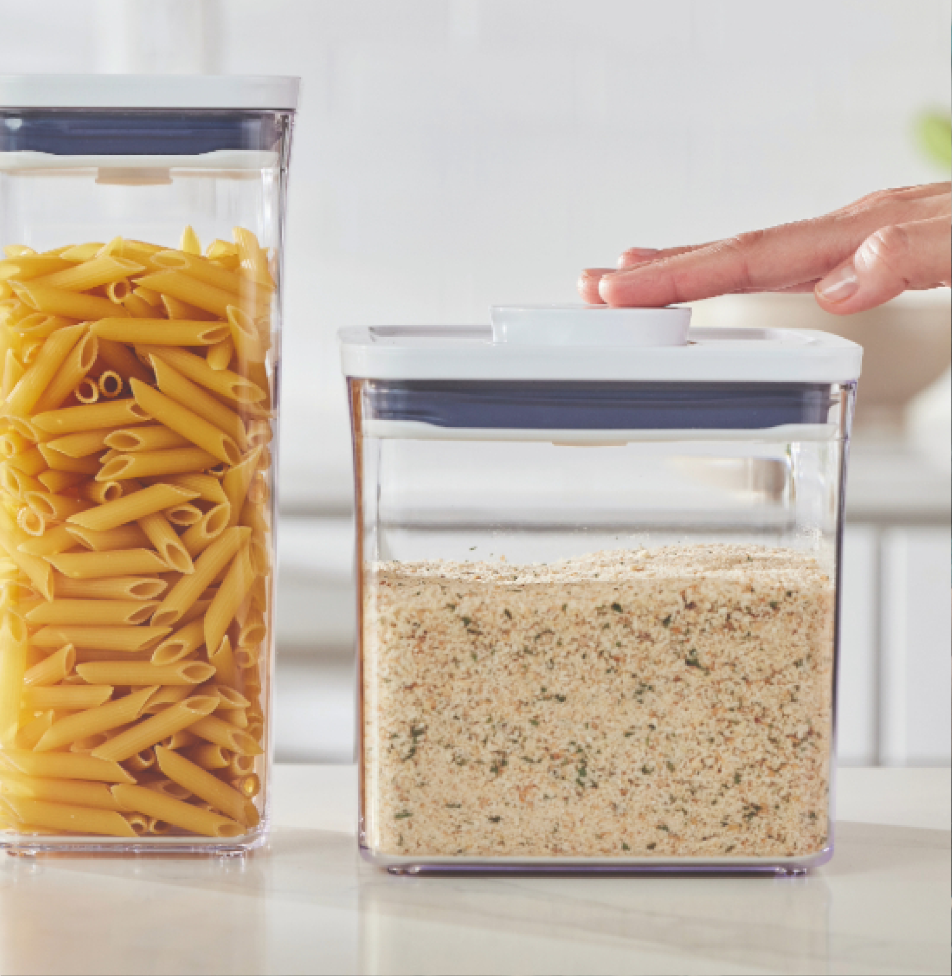 Hand pressing the lid of a clear food container beside a container filled with penne pasta.