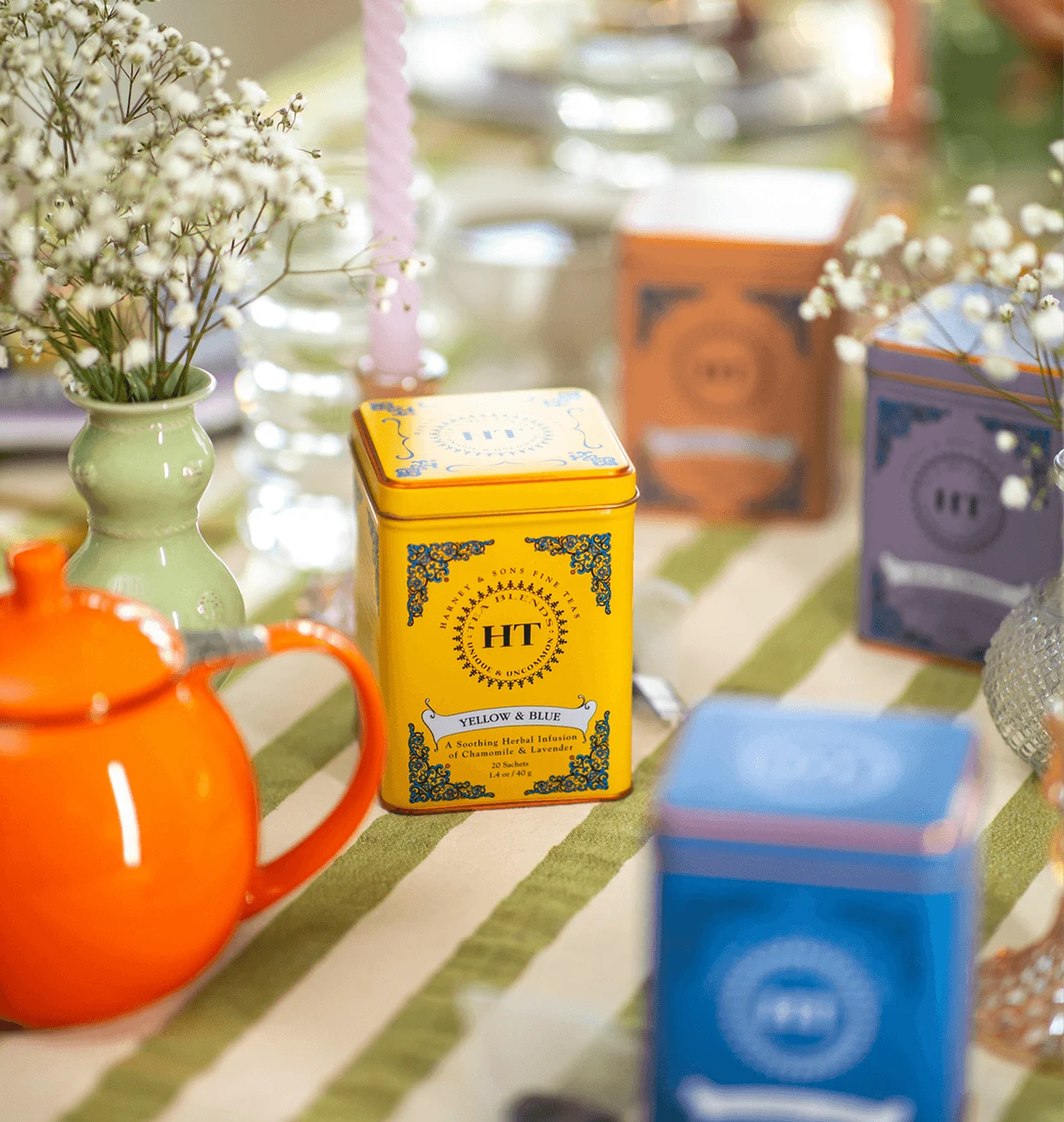 Colorful tea tins, an orange teapot, and white flowers arranged on a green-and-white striped tablecloth.