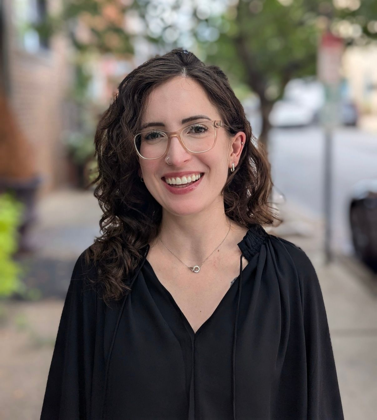 Therapist at Created Self Therapy, Gianna Damis, smiles warmly for a headshot, wearing glasses and a black top against the blurred background of a tree-lined street.