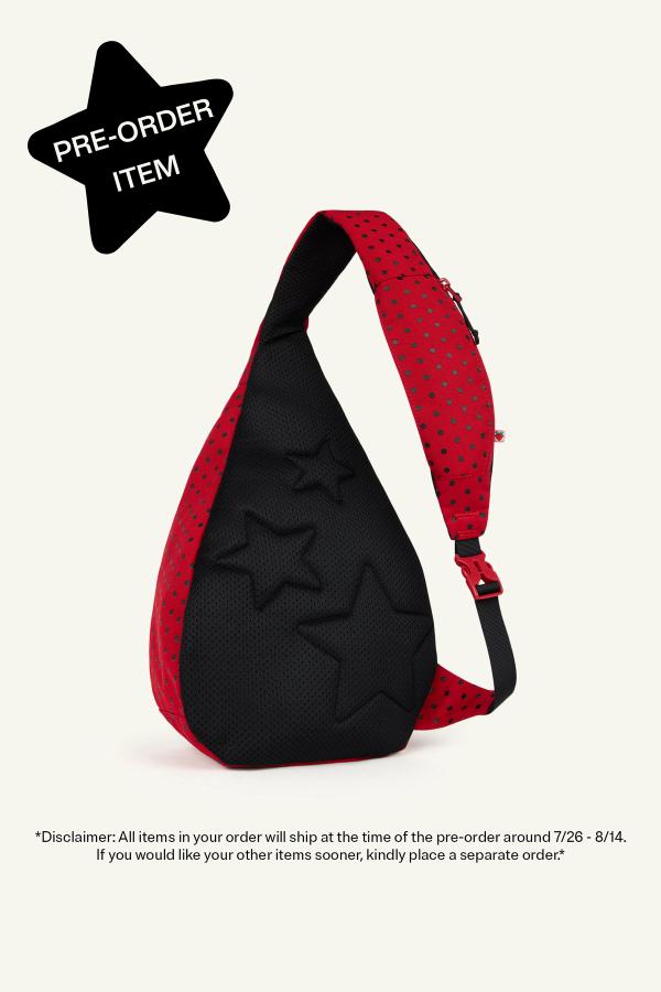 Product image of back view of a Red sling backpack with black polka dots and a black padded panel featuring embossed star shapes. The bag has a gray adjustable strap with a pink buckle and a small zip pocket on the strap. In the top left corner, a black star reads "Pre-Order Item." Bottom reads “Disclaimer: All items in your order will ship at the time of the pre-order around 7/26-8/14. If you would like your other items sooner, kindly place a separate order.