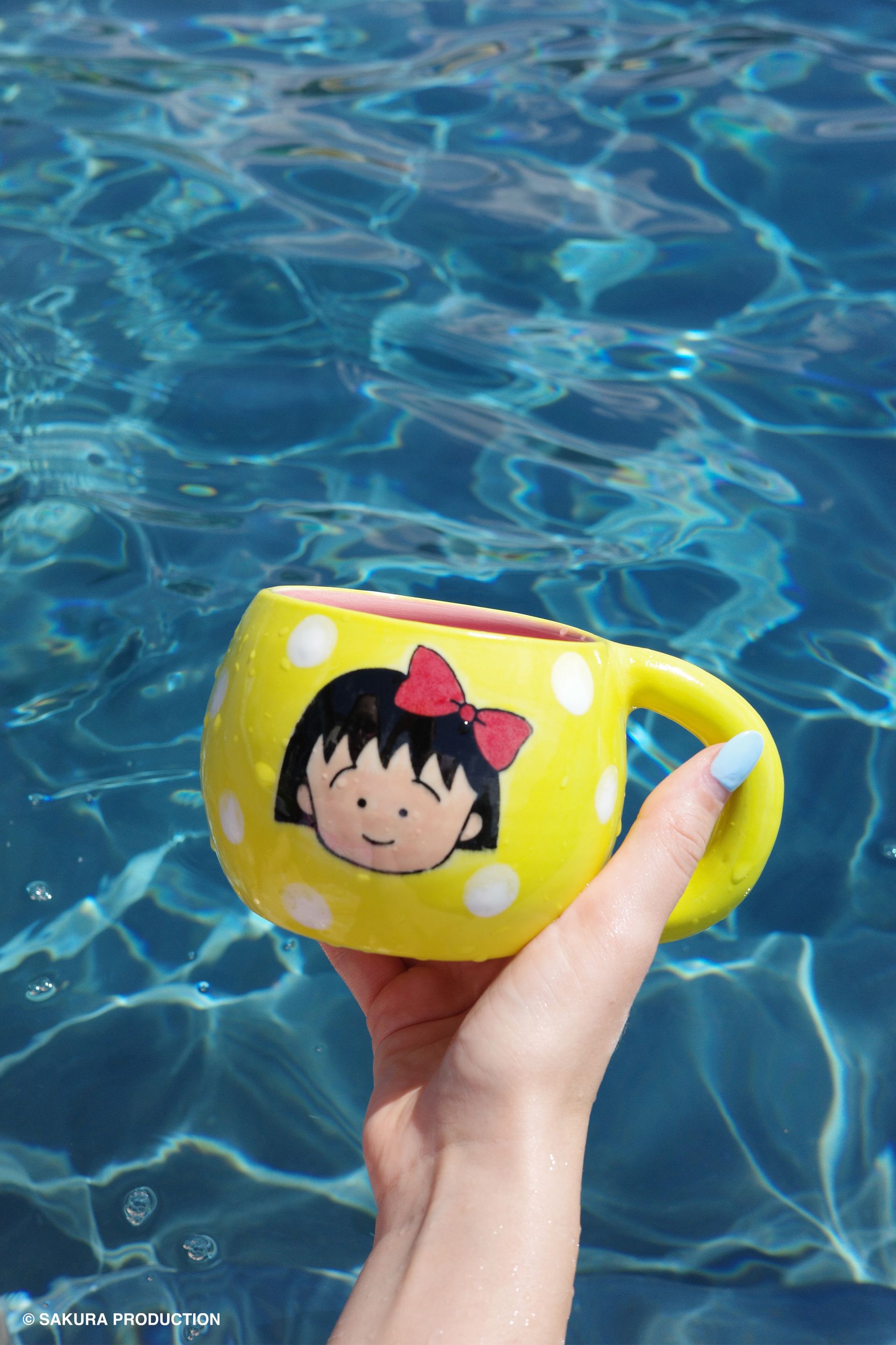 Chibi Maruko Chan hand made ceramic mug held against a pool backdrop. Yellow body and white polka dots with face of a little girl with a red bow in her hair.