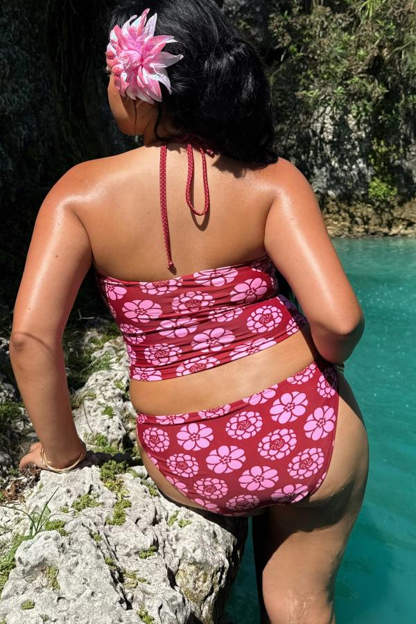 Back view of woman wearing the Kiku Print bikini bottom brief. Pattern is a maroon background with circular pink flowers. Gingham bow at the front. She stands against a body of water.
