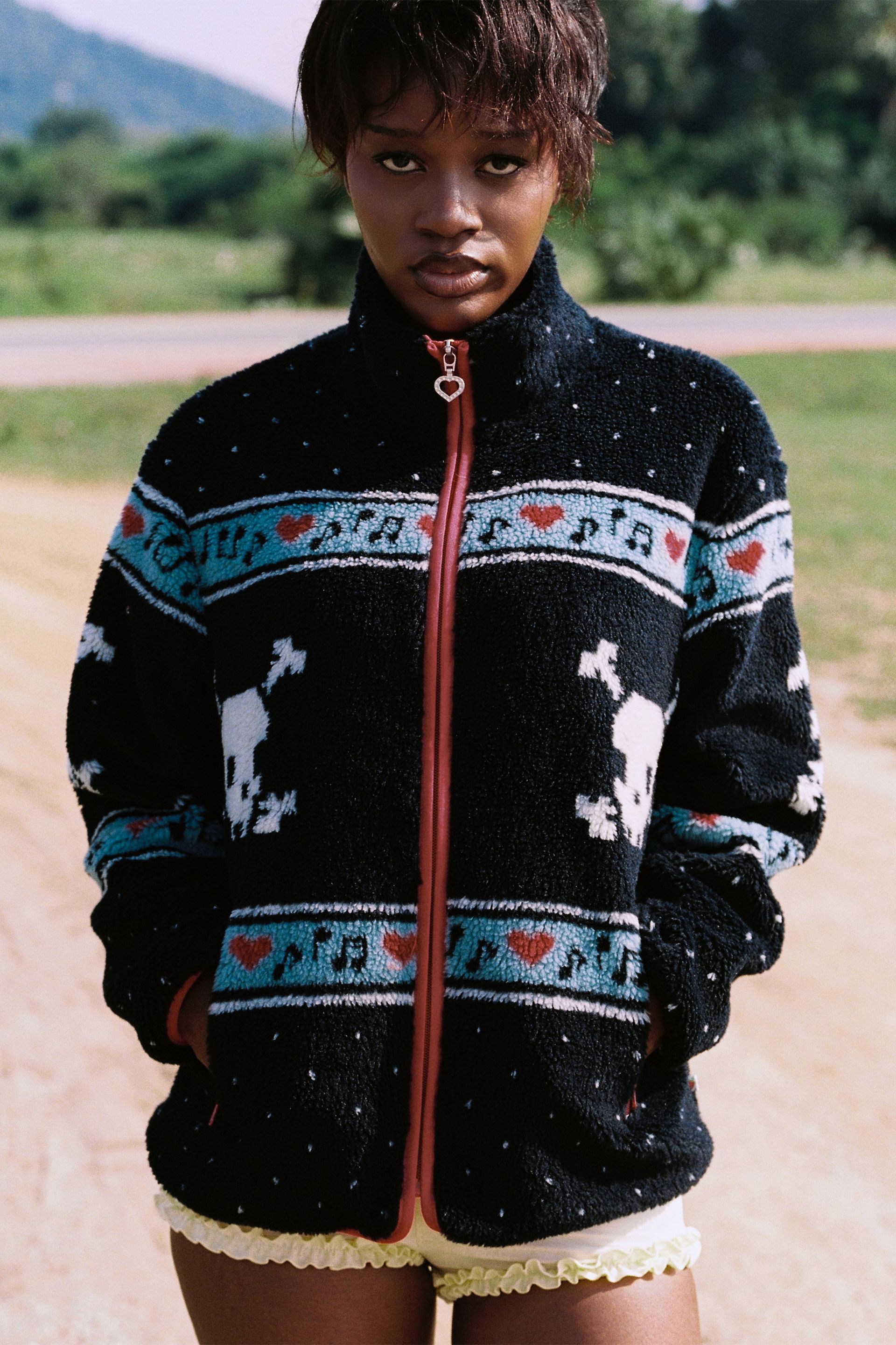 Woman wears Black Multi skully sherpa jacket featuring white skull and crossbones patterns, red hearts, and light blue musical note designs across the chest and sleeves against a natural backdrop. 