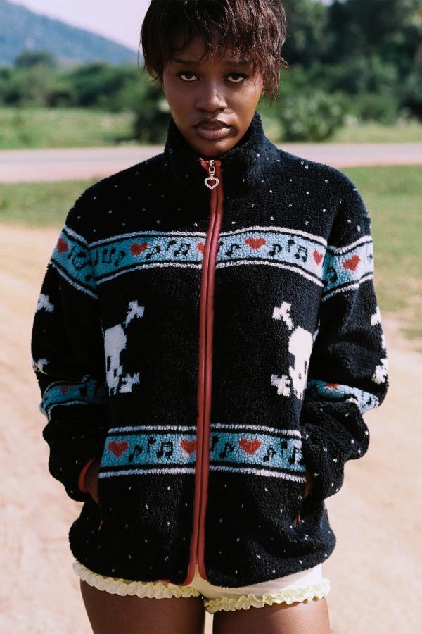 Woman wears Black Multi skully sherpa jacket featuring white skull and crossbones patterns, red hearts, and light blue musical note designs across the chest and sleeves against a natural backdrop. 