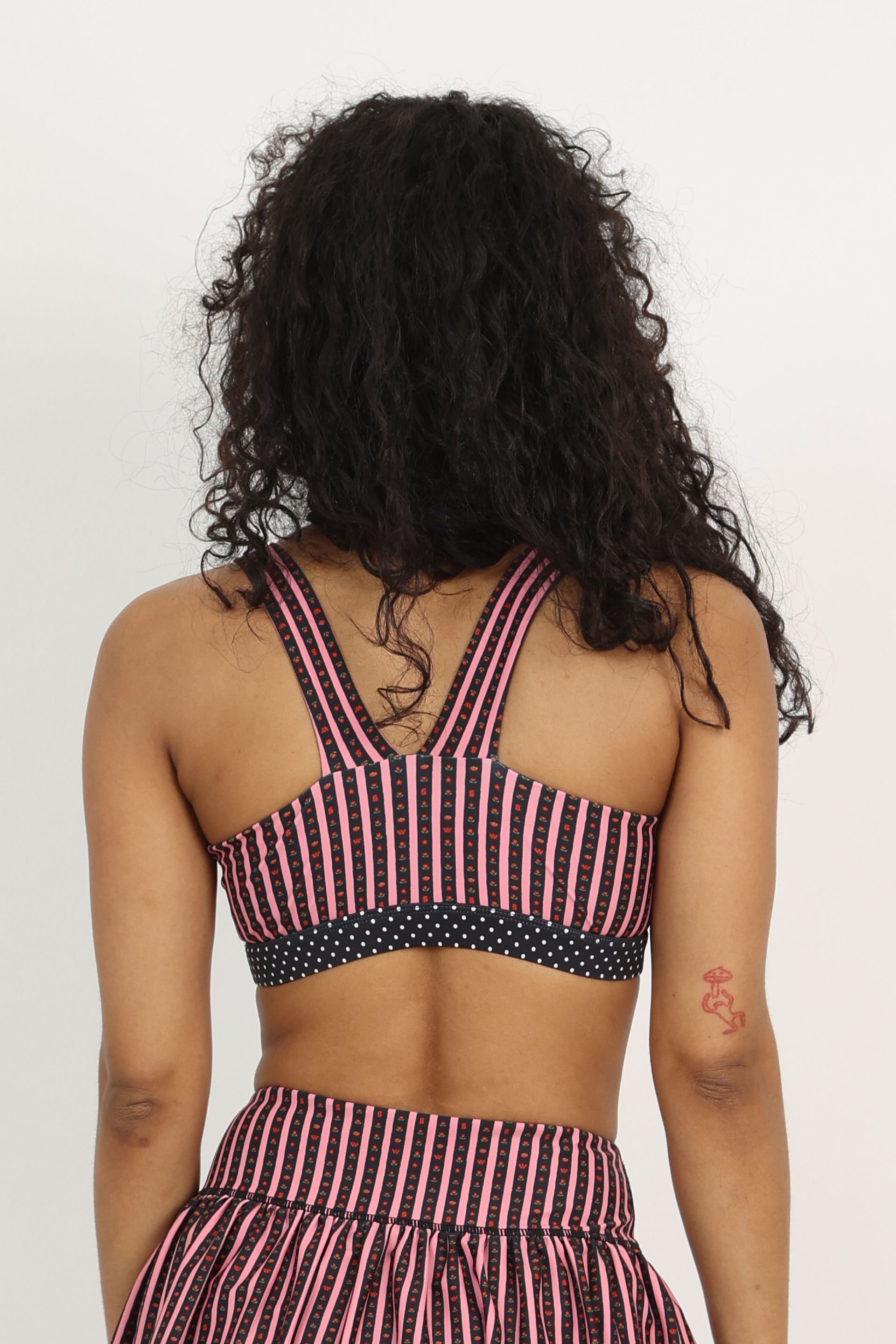 Back view of medium-support Coco Sports Bra silhouette featuring a hand-drawn, floral border stripe print  in Pink/Black with contrast polka dot print elastic band and gingham bow details.