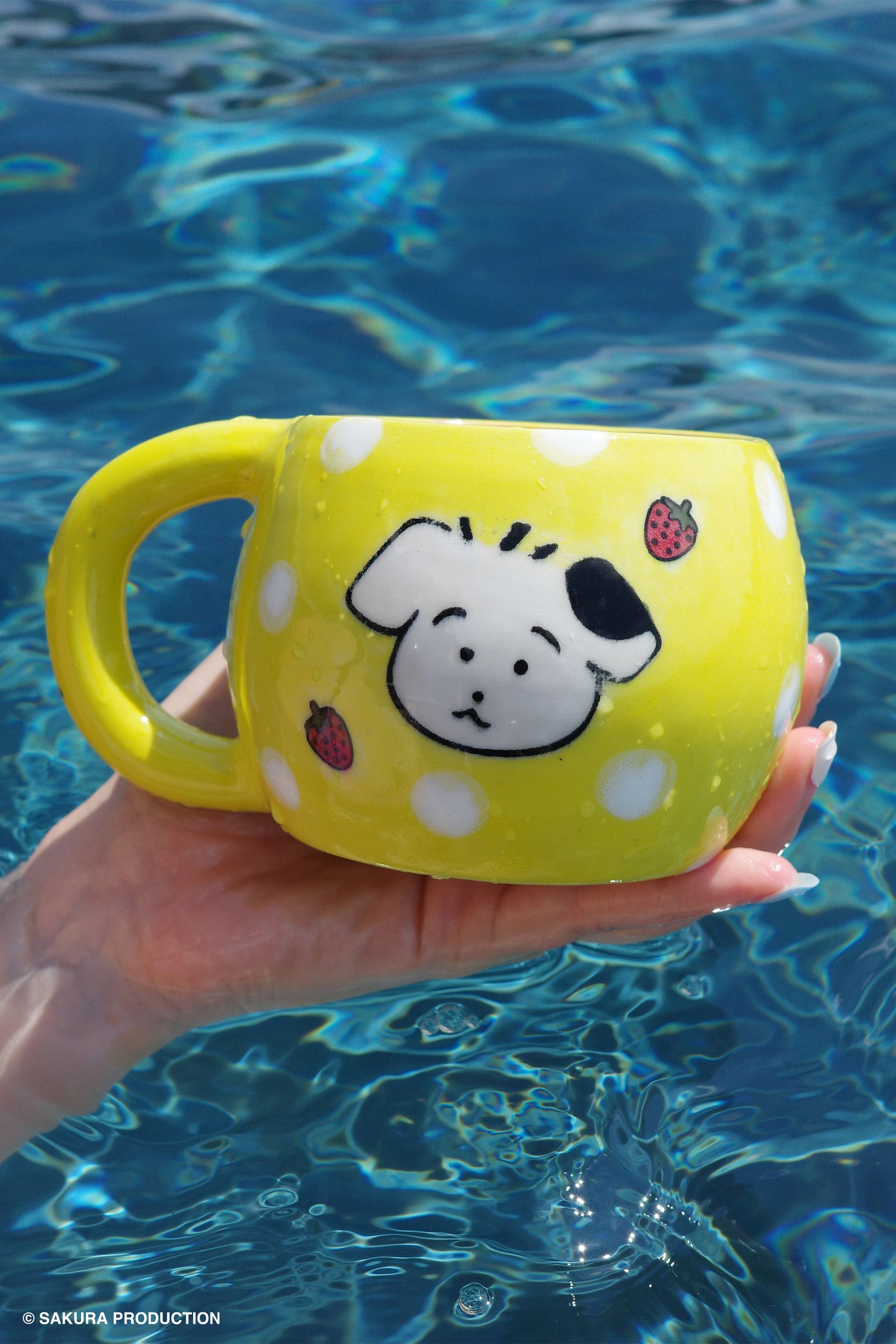 Chibi Maruko Chan hand made ceramic mug held against a pool backdrop. Yellow body and white polka dots with face of a little white and black dog surrounded by strawberries.
