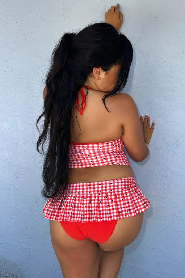 Back view of woman wearing the Red Gingham Print micro pleated skirt bikini bottom with attached red bottoms. Gingham bows at front, against a wall.
