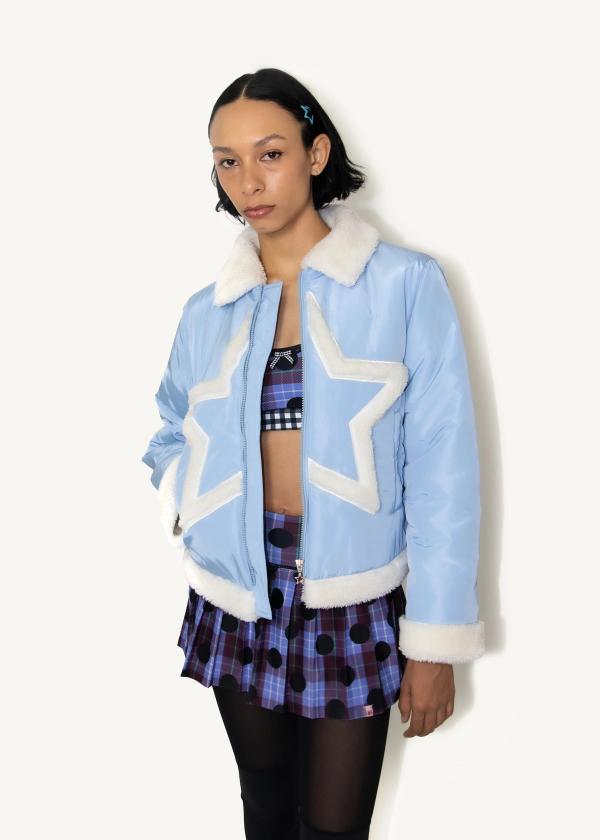 Close up of model standing with one hand in pocket of an unzipped baby blue puffer jacket with white faux fur collar, cuffs and star shape applique at front and a pleated skirt legging and sports bra underneath..